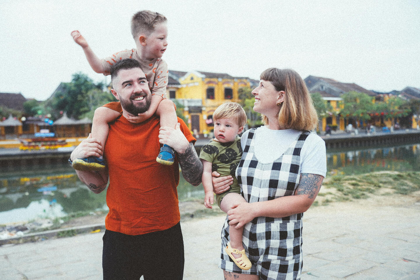 Happy family of four - father carrying young son on shoulders while mother holds toddler, all smiling together outdoors.